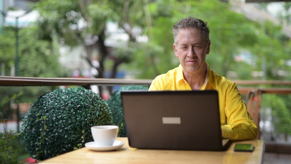 Mature Man Sitting In Coffee Shop While Using Laptop Computer And Mobile Phone