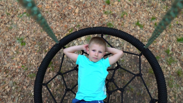 The Blue-eyed Five-year-old Boy in Blue T-shirt Lies on a Round Swing with His Hands Behind His Head