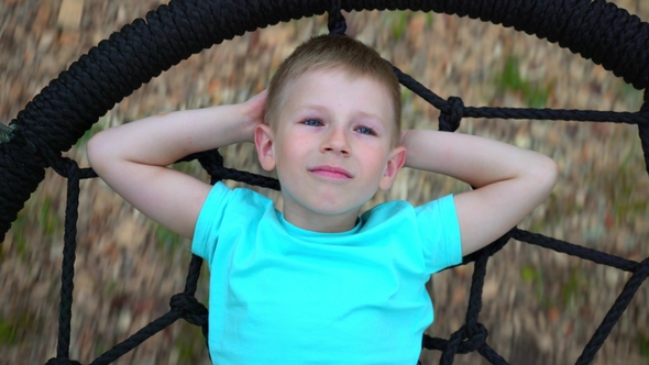 The Blue-eyed Five-year-old Boy in Blue T-shirt Lies on a Round Swing, Throws Up His Hands Behind