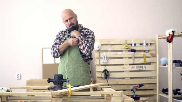 An Adult Bearded Bald Carpenter in a Green Apron Rolls Up the Sleeves of a Checkered Flannel Shirt