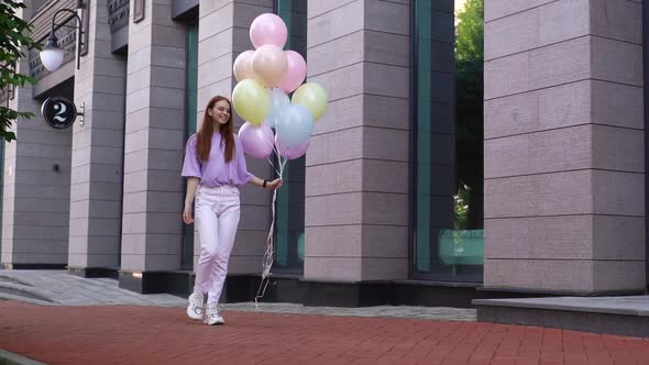 Attractive Happy Redhaired Lady Walking on City Street with Large Fountain of Colorful Balloons