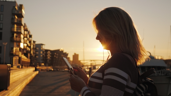 A Woman in Headphones Uses a Smartphone on the Pier on the Background of Private Yachts. At Sunset
