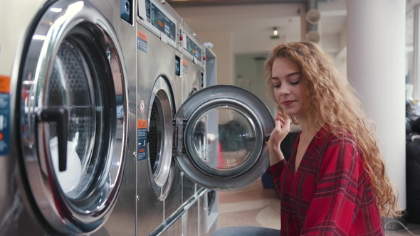 Pretty Young Woman with Red Curly Hair Put Clothes Into the Washing Mashine in the Laundry