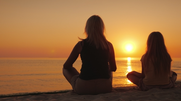 A Woman with Her Daughter Sitting Side By Side on the Sand in a Lotus Pose, Looking at the Sunset