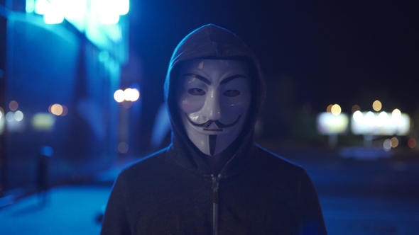 An Anonymous Hacker in a Hood Walks Down the Street and Looks at the Camera. Man with Vendetta Mask