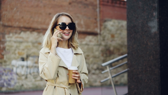 Beautiful Young Woman with Blond Hair Is Talking on Mobile Phone and Drinking Take Away Coffee