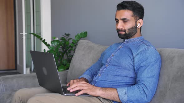 Young Adult Indian Man Typing on Laptop Keyboard Sitting on Sofa Couch