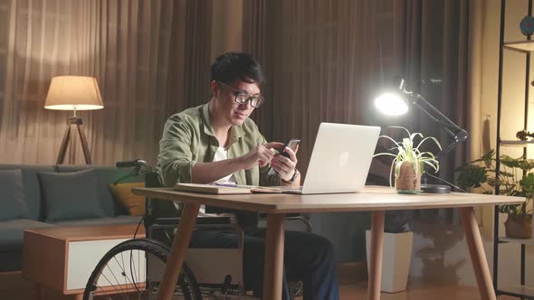 Asian Man Sitting In A Wheelchair While Working On Laptop Computer And Use Mobile Phone At Home