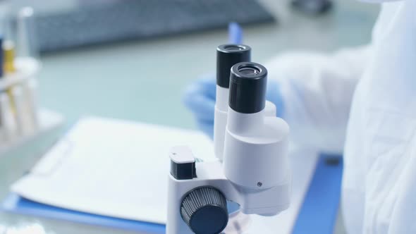 Close Up Handheld Shot of Lab Technician Taking Notes While Looking Into the Microscope