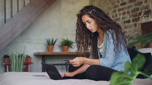 Young Woman Is Making Payment Holding Bank Card and Working with Laptop Sitting on Bed at Home