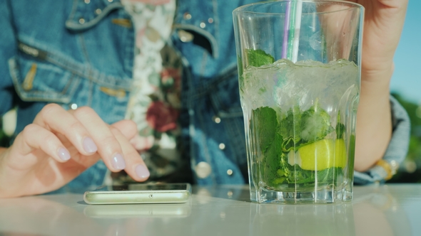 A Woman Uses a Smartphone, Next Is a Glass with a Cool Cocktail
