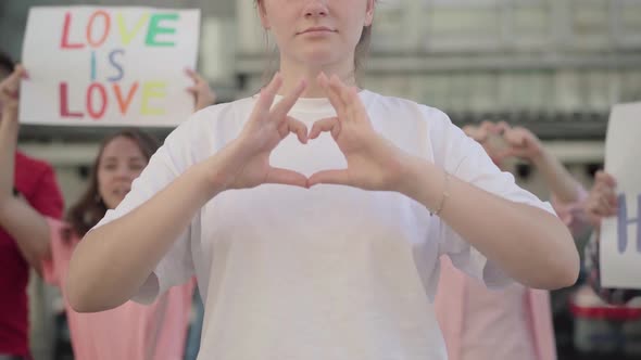 Unrecognizable Woman Making Heart with Hands As People with Anti-homophobia Banners Yelling at the