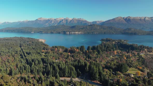 Aerial view of a forest with houses, a lake and the Andes mountains in the background.  Dronees back