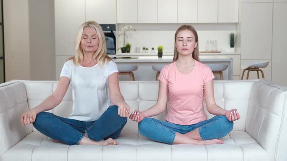 Happy Family Senior Mother and Grownup Daughter on the Sofa Practicing Yoga Meditation at Home