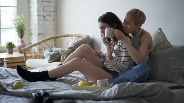 Young Happy Lesbian Couple is Drinking Coffee Sitting on Bed in Home Bedroom Spbd