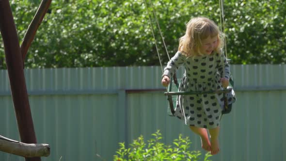 Cheerful Little Barefoot Girl in a Gray Dress is Swinging and Laughing Outdoors