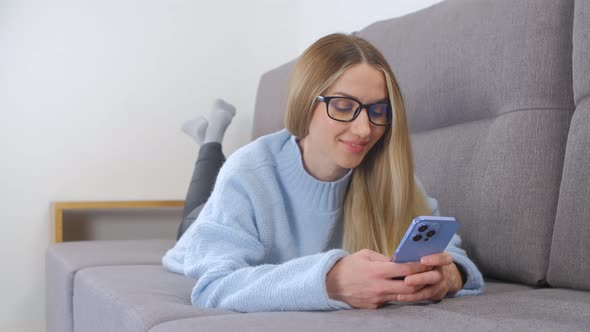 Happy blonde female using mobile phone while lying on sofa in apartment on lockdown in 4k video