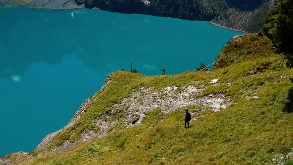 Arial shot of a blonde woman with backpack hiking down a lush mountain at a blue mountain lake in th