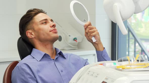 Handsome Young Man Examining His Teeth in the Mirror at the Dental Clinic