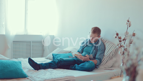 Young Man Reading Book Sitting on Sofa at Home