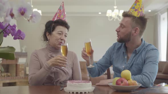 Portrait Granny and Adult Grandson Drinking Juice Sitting at the Table with Birthday Cap on Their