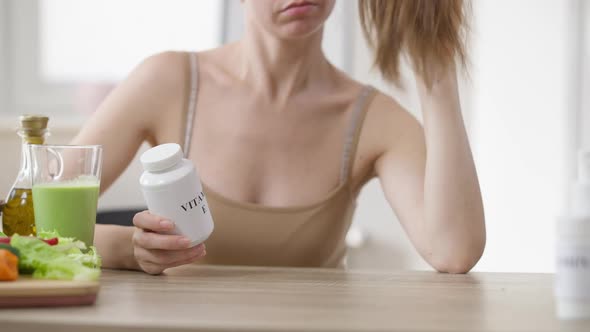 Slim Young Unrecognizable Woman Sitting at Table with Green Smoothie Holding Vitamin E Bottle