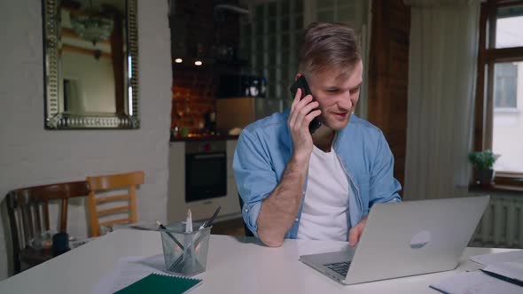 Young European Man is Talking on Phone and Using Laptop at Table in Apartment Spbas