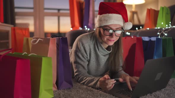 Woman with Glasses Wearing a Santa Claus Hat is Lying on the Carpet and Makes an Online Purchase