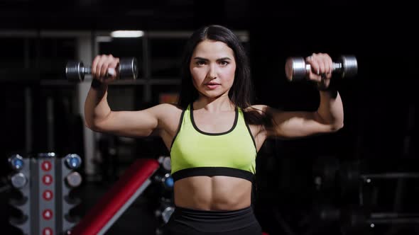 Young Woman Pumping Her Hands with Small Dumbbells Looking in the Mirror in the Gym