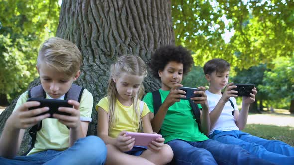 Multiracial Children Company Playing Games on Gadgets Sitting Under Tree in Park