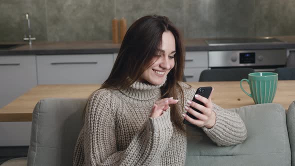 Young Caucasian Woman 25 Years Old Sits at Home on a Sofa in a Warm Sweater and Browses Quickly the