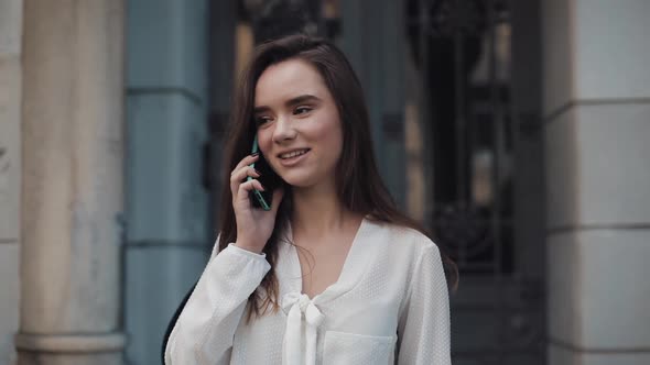 Beautiful Young Smiling Brunette Student Girl Talking on Her Smartphone, Walking Outdoors at Old