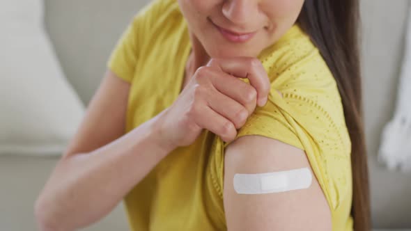 Happy asian woman sitting on sofa showing arm with plaster after vaccination