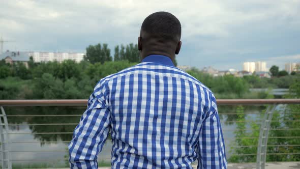 Black Man Stands on City Waterfront Near Fence in Park and Admires City View