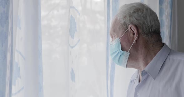 Worried Senior Man in Medical Mask Looks Thoughtfully at Window During Isolation