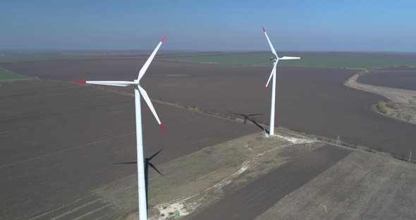 Aerial drone footage of two rotating wind turbines in plowed fields during spring