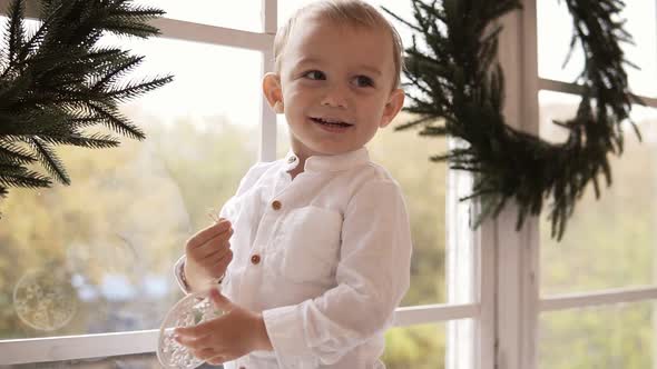 Adorable Blonde Toddler in White Shirt Standing on the Window Sill Holding a Christmas Toy and