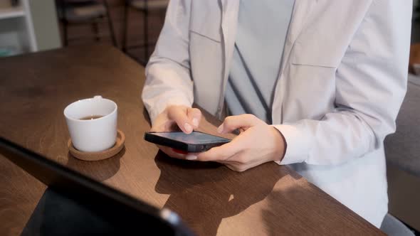 Female hands click the phone in the morning in the kitchen. Girl sitting at the table drinking tea