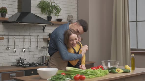 Young happy husband hug smiling wife cutting vegetable for salad in kitchen