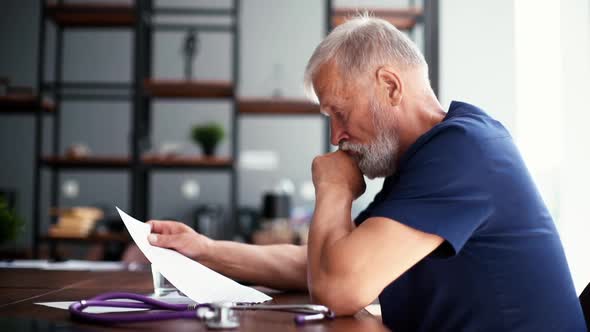 Concentrated old gray-haired mature male doctor reading medical report of patient sitting at desk.