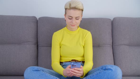 Young white female using mobile phone for communication online while sitting on sofa at home