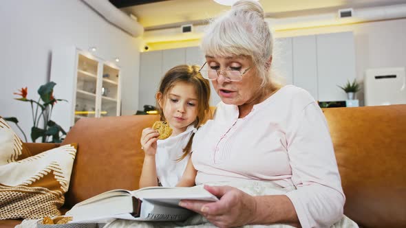 Grandmother is Reading a Book to Her Little Girl Eating Homemade Cookie While Sitting on Sofa Studio