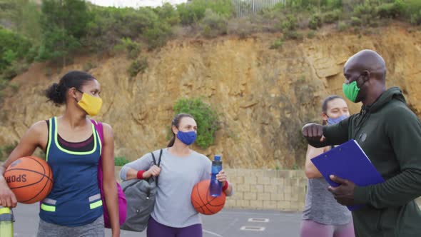Diverse female basketball team and male coach wearing face masks greeting with elbows
