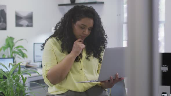 Thoughtful biracial businesswoman working, using laptop holding her chin in modern office