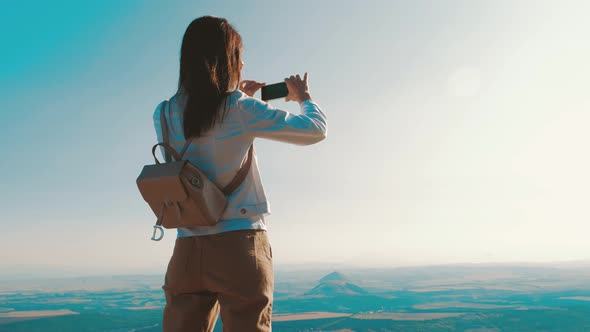 Young Woman Traveler on a Background of Mountains Takes Photos on a Mobile Phone