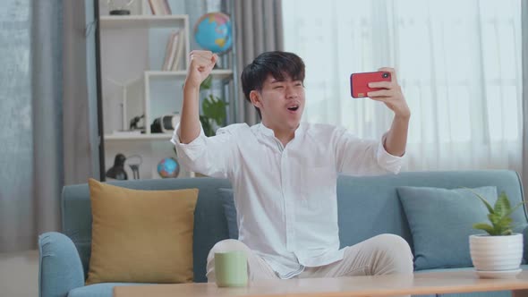 Asian Man Celebrating Winning Game On Smartphone While Sitting On Sofa In The Living Room