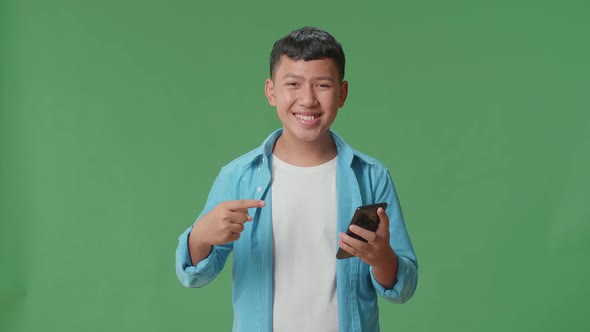 Smiling Young Asian Boy Use Phone And Pointing To Smart Phone In The Green Screen Studio