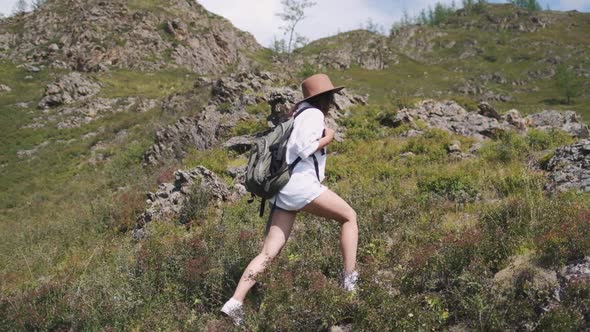 Attractive Brunette Girl in a Hat with a Backpack Travels in the Mountains. Woman in a White Shirt