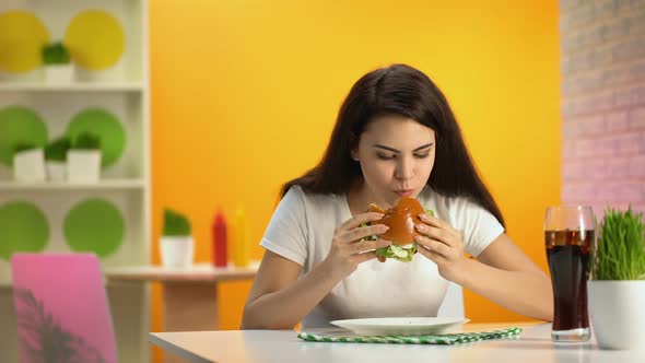 Hungry Young Lady Eating Tasty Cheeseburger in Cafe, Soft Drink Glass on Table