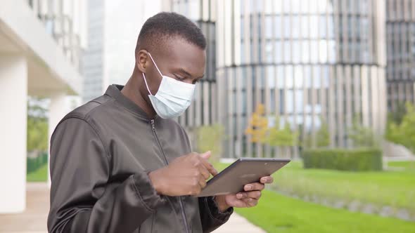 A Young Black Man in a Face Mask Works on a Tablet - Office Buildings in the Blurry Background